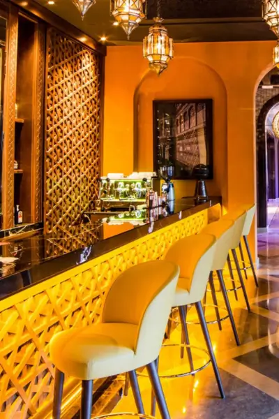Stylish bar counter at Quattro Marrakech featuring yellow upholstered stools, wooden lattice screens, and hanging Moroccan lanterns