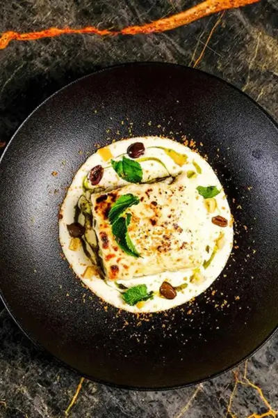Square cut lasagna in a creamy white sauce with olives and fresh mint garnish on a black plate at Quattro restaurant, Marrakech